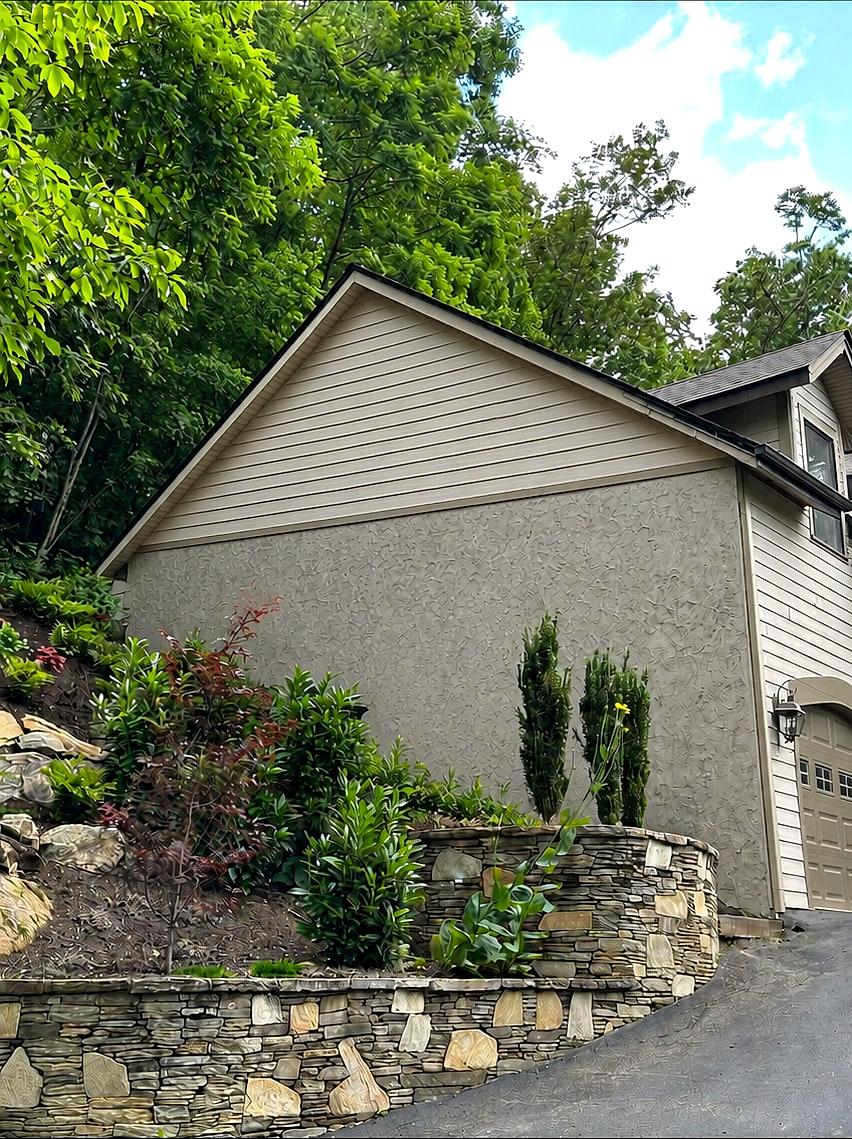 The same hillside elevation before the addition — a blank stucco gable end above the stacked-stone terraces, with no porch or deck