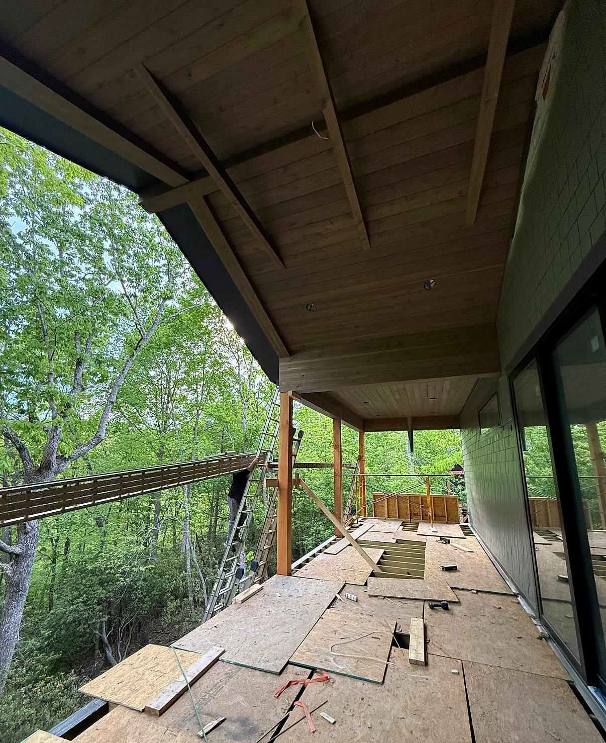 Covered deck under construction — stained tongue-and-groove soffit and exposed rafters above a subfloor still being laid, out over a wooded slope